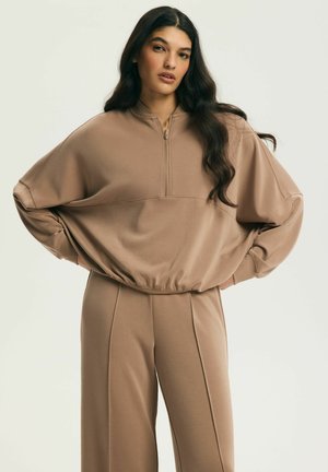 Kobieta o długich ciemnych włosach, ubrana w luźną beżową bluzę na zamek oraz pasujące szerokie spodnie, stojąca z rękami na biodrach.