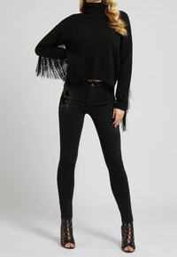 Femme portant un pull à col roulé noir avec des manches à franges, un jean skinny noir orné d'embellissements, et des talons ouverts à lacets noirs.