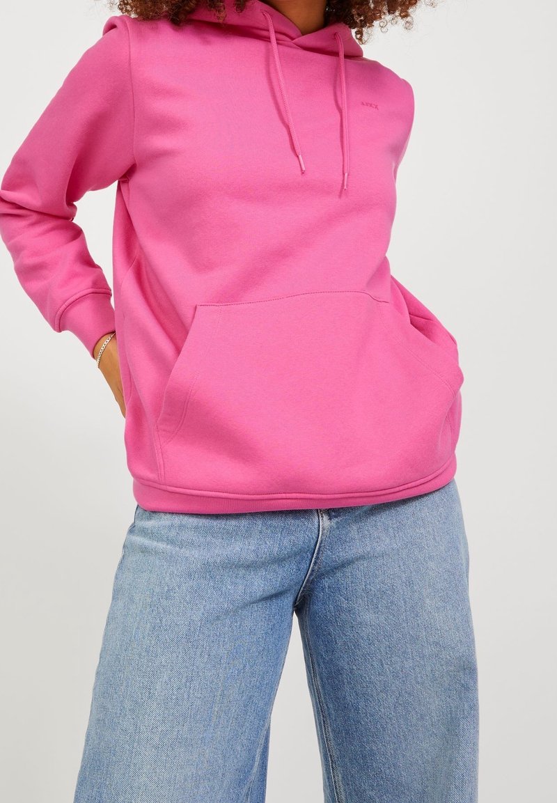 Personne portant un sweat à capuche rose vif avec une poche avant et un jean en denim bleu clair sur un fond uni.