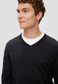 Czarny sweter z dekoltem w serek wykonany z dzianiny, z subtelnym logo na piersi, nałożony na biały T-shirt.