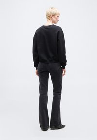 Άτομο με ξανθά κοντά μαλλιά που φοράει μαύρο φούτερ και σκουρόχρωμο τζιν με καμπάνα, στέκεται γυρισμένο προς τα πίσω μπροστά σε ένα απλό ανοιχτό φόντο.