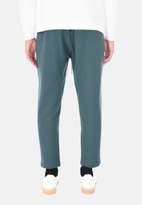 Vista posteriore di una persona che indossa pantaloni dritti teal, calzini neri, sneakers bianche e una maglietta a maniche lunghe bianca su uno sfondo semplice.