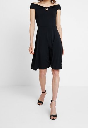 Kvinde iført en sort knælængde kjole med bar skuldre og hjerteformet udskæring samt sorte hæle med ankelsnor, stående mod en hvid baggrund.