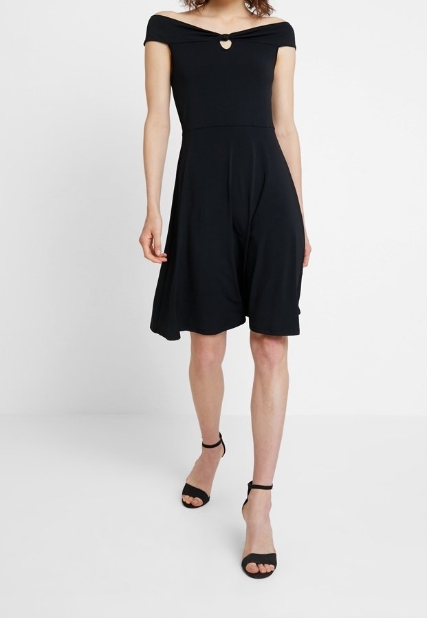 Femme portant une robe noire mi-longue à épaules dénudées avec une découpe en forme de cœur et des talons noirs à bride cheville, debout devant un fond blanc.