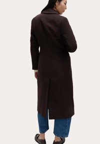 Brązowy płaszcz z mieszanki wełny o dopasowanym kroju, z klapami na kołnierzu i rozcięciem z tyłu. Noszony na niebieskie, krótkie jeansy i czarne płaskie buty.