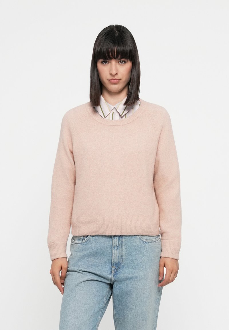 Maglione a maglia morbida di un delicato rosa chiaro, con collo rotondo e orlo a costine. Indossato sopra una camicia con colletto a righe verticali. Fit casual.