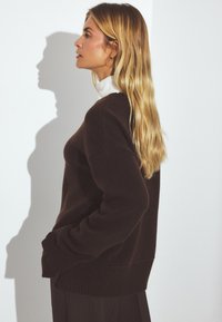 Pull en tricot marron avec une coupe décontractée, ourlet et poignets côtelés, porté par-dessus un col roulé blanc. Cheveux longs et ondulés visibles.