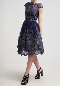 Tummansininen mekko, jossa on hihattomat hihat, monimutkainen pitsikuvaus, kukkakuviot ja flare-helma. Yhdistetty mustiin korkokengiin.