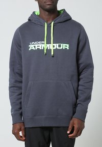 Sudadera gris oscuro con un bolsillo frontal, cordones contrastantes de color verde neón y el logo de "Under Armour" en blanco bold. Tejido suave y corte relajado.
