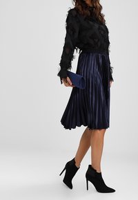 Jupe plissée marine avec un pull noir transparent à franges texturées, associé à des bottines noires à talons hauts et une pochette bleue.