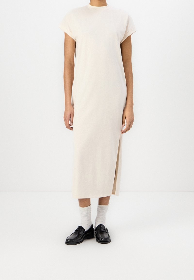 Personne debout portant une robe midi beige sans manches avec une fente latérale, des chaussettes blanches et des mocassins noirs sur un fond blanc uni.