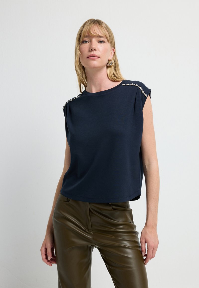 Himmelblaues T-Shirt mit kurzen Ärmeln und lockerem Schnitt, ausgestattet mit dekorativen silbernen Nieten auf den Schultern und einem glatten, leichten Stoff.