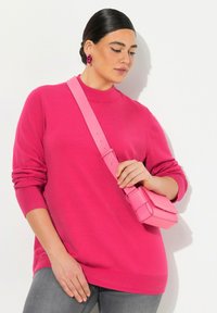 Kuumapinkki villapaita korkealla kauluksella ja pitkillä hihoilla, yhdistettynä pieneen vaaleanpunaiseseen olkalaukkuun. Mallilla on päällään harmaat farkut ja rohkeat korvakorut.