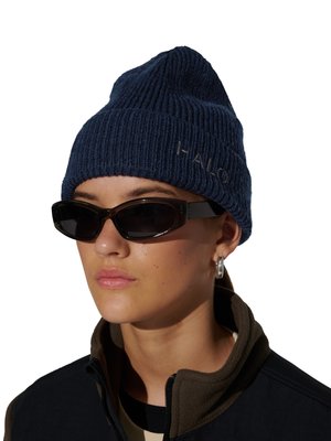 Person iført en marineblå strikket hue med teksten "HALO", sorte smalle solbriller, sølv hoops-ørering og en mørk lynlåstrøje med brun krave.