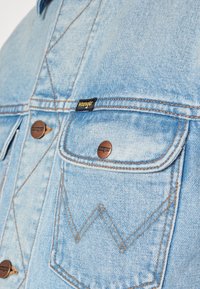 Jasnoniebieska kurtka dżinsowa z miedzianymi elementami, z dwoma kieszeniami na piersi, kontrastowym przeszyciem i metką z logo "Wrangler".