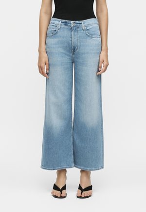 Γυναίκα που φοράει ανοιχτό μπλε cropped τζιν με φαρδιά μπατζάκια, μαύρο αμάνικο τοπ και μαύρα σανδάλια ανοιχτής μύτης, στέκεται μπροστά σε λευκό φόντο.