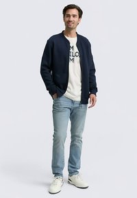 Marinblå bomberjacka med ribbad krage, kombinerad med en ljusfärgad t-shirt med tryck, ljusblå jeans och vita sneakers.