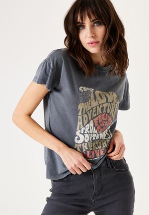 T-shirt en coton gris à manches courtes avec un graphique de texte coloré. Coupe décontractée avec col rond, porté avec un jean noir.