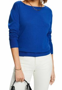 Blauer Strickpullover mit langen Ärmeln und U-Boot-Ausschnitt. Getragen mit weißer Hose; hält eine schwarze Tasche in einer Hand. Glatte Textur.