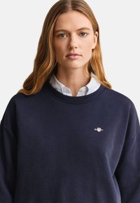 Tânără cu păr lung castaniu deschis poartă o cămașă albă cu guler sub un hanorac bleumarin cu un mic logo brodat pe piept.