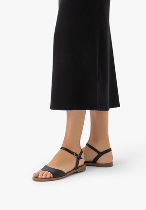 Fekete lapos szandált viselő lábak bokapánttal és csatokkal, középhosszú fekete szoknyával, fehér háttér előtt.