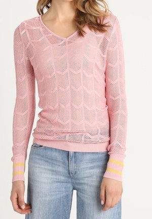 Kobieta ubrana w różowy sweter z długim rękawem wykonany na drutach z wzorem w jodełkę i żółtymi paskami na mankietach, w połączeniu z jasnoniebieskimi dżinsami.