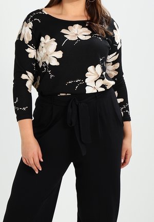 Femme portant un haut noir à manches longues avec un motif floral beige et un pantalon noir taille haute avec ceinture à nouer, debout devant un fond uni.