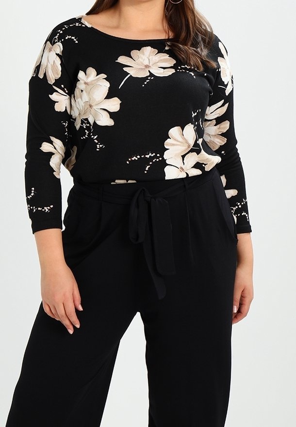 Femme portant un haut noir à manches longues avec un motif floral beige et un pantalon noir taille haute avec ceinture à nouer, debout devant un fond uni.