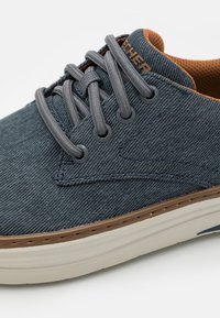 Sneaker in denim blu con lacci grigi, dettagli marroni sulla linguetta e una texture a motivo. Presenta una suola bianca ammortizzata e occhielli metallici.