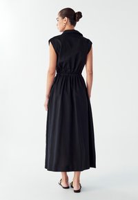 Juoda be rankovių maksi suknelė su apykakle, raišteliu liemeniui ir glotnia tekstūra. Pasižymi ilgu, sklandžiu dizainu ir minimalistiniu stiliumi.