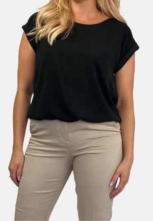 Femme portant un haut noir ample à manches courtes et un pantalon beige ajusté, debout contre un fond clair uni.