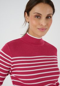Donna con carnagione chiara che indossa un maglione rosa a coste con strisce orizzontali bianche, che guarda leggermente fuori campo con un sorriso delicato.