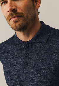 Marineblauwe gebreide polo met een gestructureerde stof, drie knoppen en een klassiek kraagontwerp. Close-up van de halslijn en knoppen.