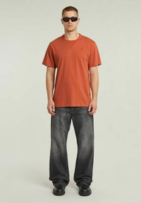 T-shirt di cotone color ruggine con maniche corte, abbinato a jeans neri in denim strappati. Il modello indossa occhiali da sole neri e calzature nere.