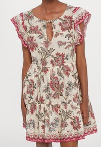 Robe à imprimé floral couleur beige clair avec des manches volantées, un décolleté en trou de serrure et une jupe à volants. Des motifs en rouge, noir et vert ornent le tissu.