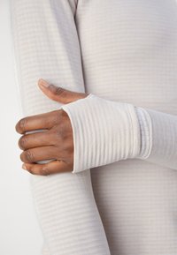 T-shirt à manches longues en crème clair avec des fines rayures verticales. Le tissu est doux et extensible, avec des poignets ajustés dotés de trous pour les pouces.
