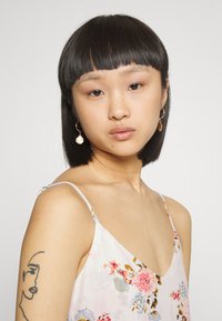 Vestido florido, sem mangas, com um design em V em tecido leve. Apresenta uma tatuagem delicada no braço e brincos de argola dourados com detalhes.