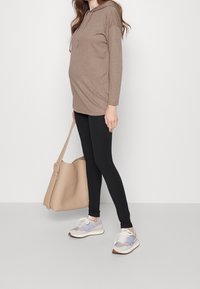 Felpa a maniche lunghe marrone con costine e cappuccio, abbinata a leggings neri e sneakers grigie con dettagli blu. Porta una borsa beige.