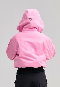 Személy élénk rózsaszín kapucnis dzsekiben és fekete nadrágban, hátulról nézve, egyszerű világos háttér előtt.