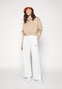 Beige cropped sweatshirt med krave og logo, kombinert med løstsittende hvite joggebukser med sidesplitter og logo. Hvite sneakers og en oransjeaktig bøttehatt fullfører antrekket.