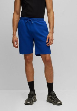 Shorts de sport bleu en tissu doux, dotés d'une taille élastique avec cordon de serrage, et d'une coupe décontractée. Associés à des baskets noires.