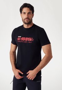 Černé bavlněné tričko s výrazným červeným grafickým potiskem s nápisem "GUESS 1981 ATHLETIC DEPT. CALI." Navrženo s klasickým kulatým výstřihem.