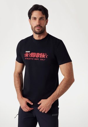 Juodas medvilninis marškinėlis su ryškiu raudonu grafikiniu spaudiniu, kuriame parašyta "GUESS 1981 ATHLETIC DEPT. CALI." Sukurtas su klasikinio apvalaus kaklo iškirpimu.