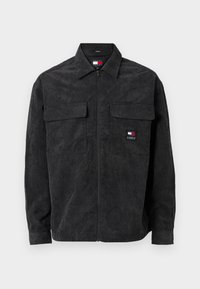 Musta corduroy-takki, jossa on etuvetoketju, kaksi rinta-taskua ja pieni logo vasemmalla puolella. Takissa on kauluspäällinen ja ribbauskaitaleet.