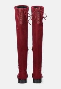 Botas altas de ante rojo con detalles de cordones en la parte trasera, cremallera lateral y tacones bajos negros, mostradas desde la parte trasera sobre un fondo liso.
