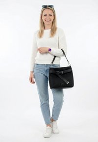 Naeratav naine valge kampsuni ja teksadega, kandmas õlal musta käekotti, seljas valged tossud ja lilla käekell, peal päikeseprillid.