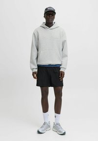 Person in einem grauen Hoodie, schwarzen Shorts, weißen Socken, silbernen Sneakers und einer dunklen Mütze, der vor einem schlichten weißen Hintergrund steht.