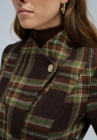 Abrigo de lana de cuadros marrón con acentos en verde y blanco, que presenta un solapa diagonal y detalles de botones en tono dorado. Tejido ligero, corte entallado.