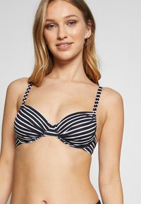 Bikini vrch s čierno-bielymi pruhmi, s podpornou výstužou, nastaviteľnými ramienkami a prekrúteným detailom na prednej strane. Hladký textilný materiál.