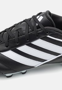 Botín de fútbol negro con rayas diagonales blancas, hecho de cuero sintético. Presenta una superficie texturizada, diseño de cordones y tacos moldeados en la suela.